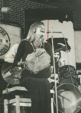 Rita Lee seated, playing a percussion instrument and singing with the group Os Mutantes.