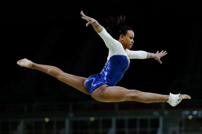 Den brasilianske idrettsutøveren Rebeca Andrade, i hvit badedrakt, gjør et hopp med bena og armene strukket i været.