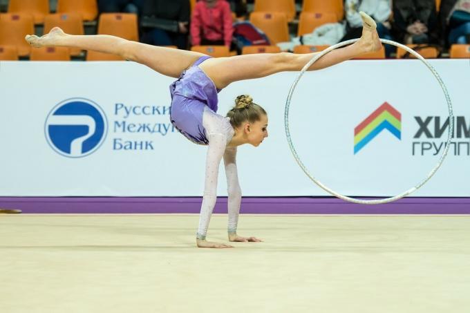 Rytmisk gymnast støtter seg med armene strukket ut på gulvet og bena åpne oppover, holder bøylen med en fot.