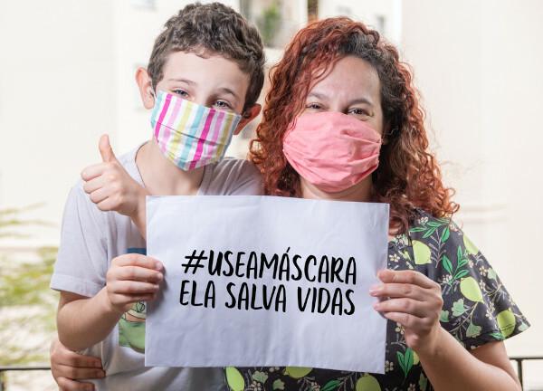 En kvinde og en lille dreng, iført ansigtsmasker, holder et skilt sammen, hvor der står: "#bær en maske — det redder liv"