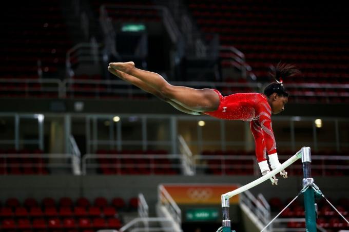 Gymnast Simone Biles, in a red bathing suit, jumps from one uneven bar to another.