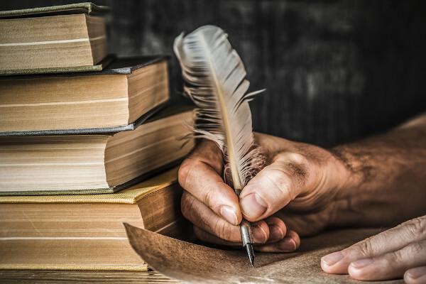 Nærbillede af en persons hånd, der skriver et brev med en fjerpen, ved siden af ​​flere gamle bøger.